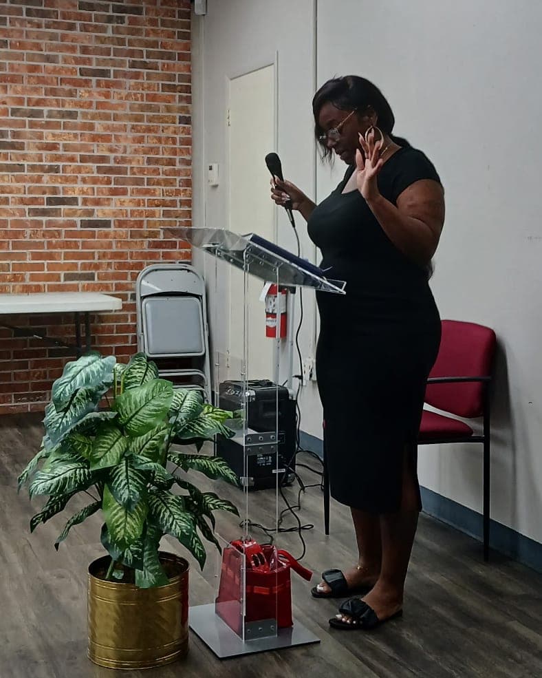 A woman preaches from a podium, microphone in one hand and the other hand raised.